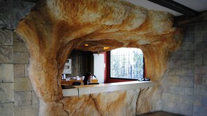 A stone-walled room with a large rock archway, a wooden reception desk, and a window with a view of trees. - Pirates Village (Calvia)