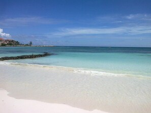 Ein Strand mit klarem türkisfarbenem Wasser, einem steinernen Wellenbrecher und einer in der Ferne liegenden Küste mit Gebäuden.