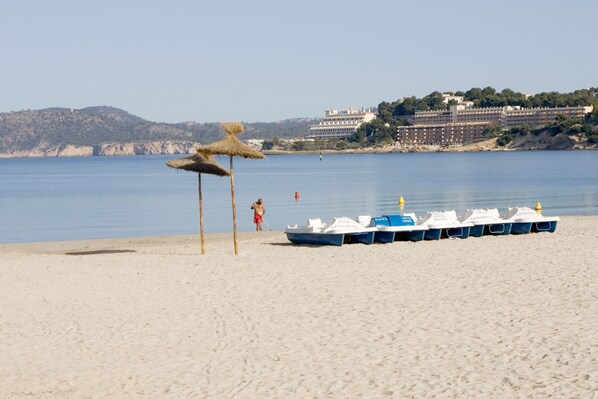 A beach with a row of boats, a thatched umbrella, and a building in the background. - Plazamar Serenity Resort (Calvia)