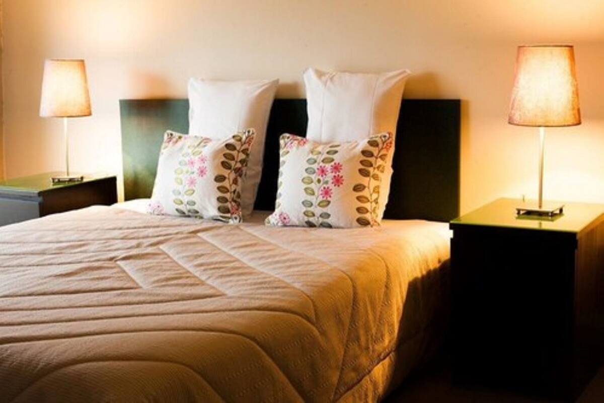 Ein ordentlich gemachtes Bett mit gesteppter Bettdecke und zwei dekorativen Kissen mit Blumenmuster. Auf beiden Seiten des Bettes stehen je eine Nachttischlampe.