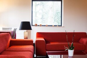 Un salon moderne avec un canapé rouge, une petite table, une lampe et une fenêtre donnant sur des arbres.
