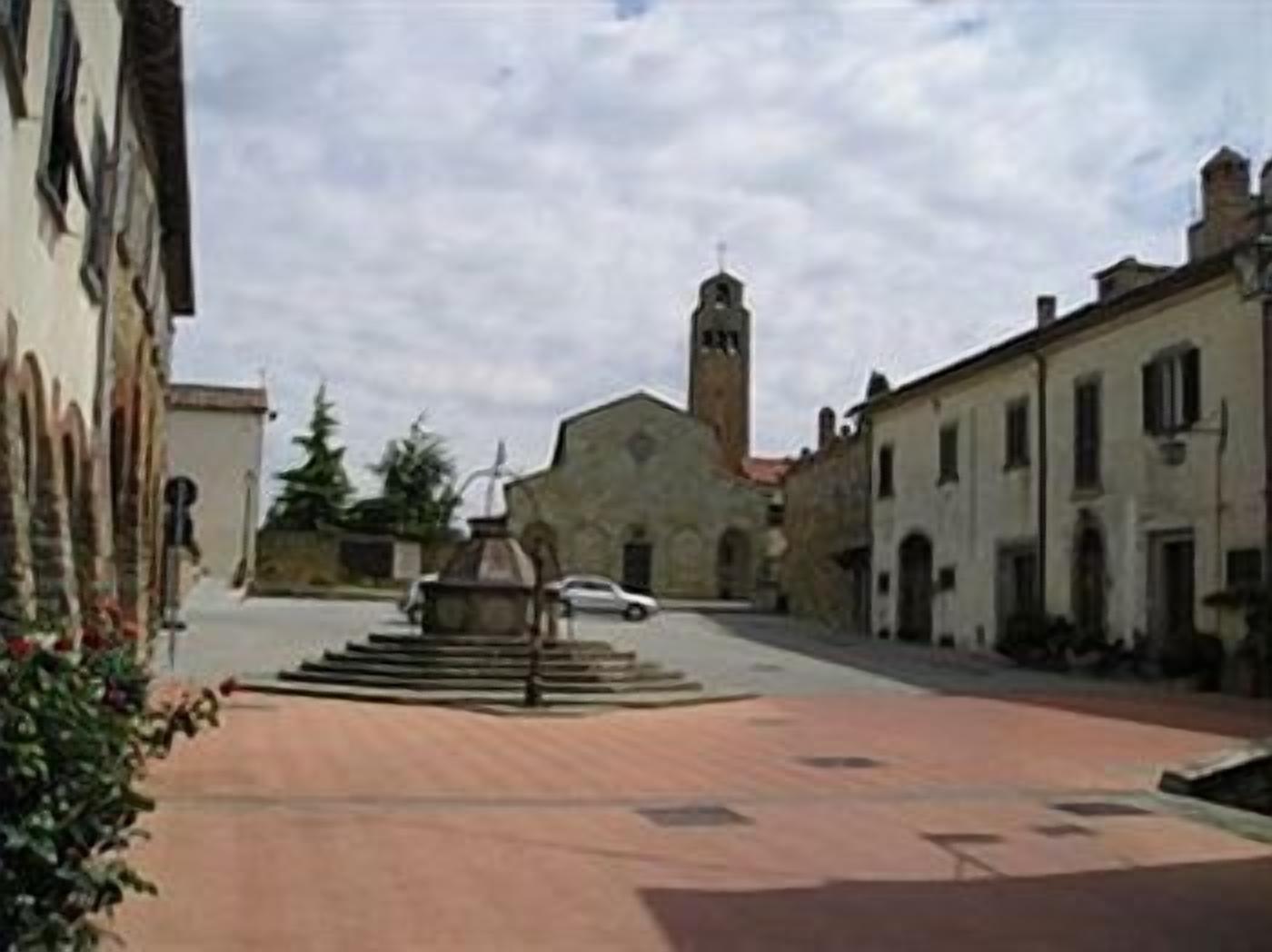 Eine ruhige Straße mit einem Brunnen in der Mitte, umgeben von historischen Gebäuden, im Hintergrund ein Kirchturm.