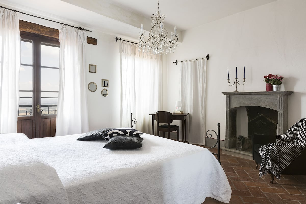 A bedroom with a large bed, a chandelier, a fireplace, and a view through a window.