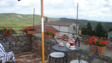 Eine Terrasse mit gedecktem Tisch für zwei Personen, umgeben von roten Blütenpflanzen in Töpfen, mit Blick auf eine Hügellandschaft.