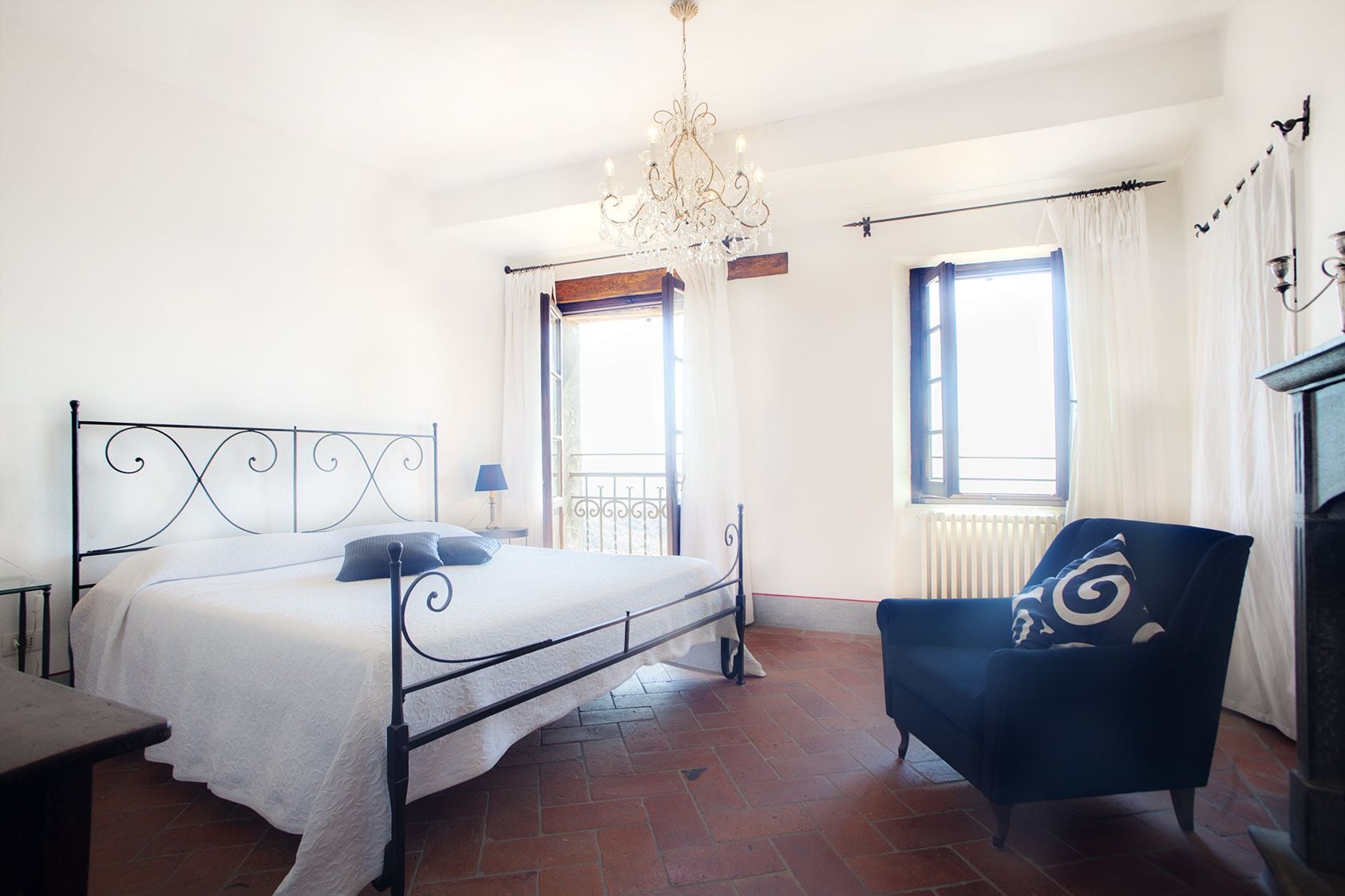 A bedroom with a bed, a chair, a chandelier, and two windows.