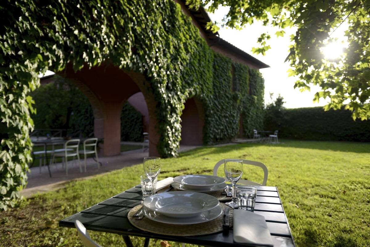 Une table dressée à l’extérieur, avec des assiettes, des verres et des couverts, placée sous une structure recouverte de lierre.
