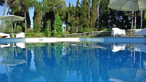 Una piscina cristalina con sillones de descanso blancos y sombrillas, rodeada de altos árboles.