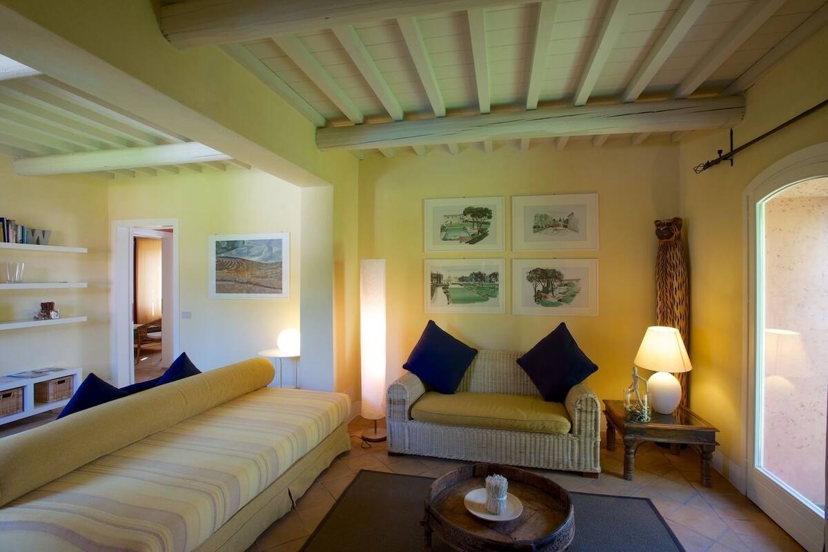 Un salon chaleureux avec un canapé, un fauteuil en osier et une table basse. Des tableaux encadrés ornent le mur, et une lampe est posée sur une table d’appoint.