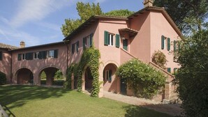 Una villa rosa con paseos en arco y contraventanas verdes, rodeada de abundante vegetación.