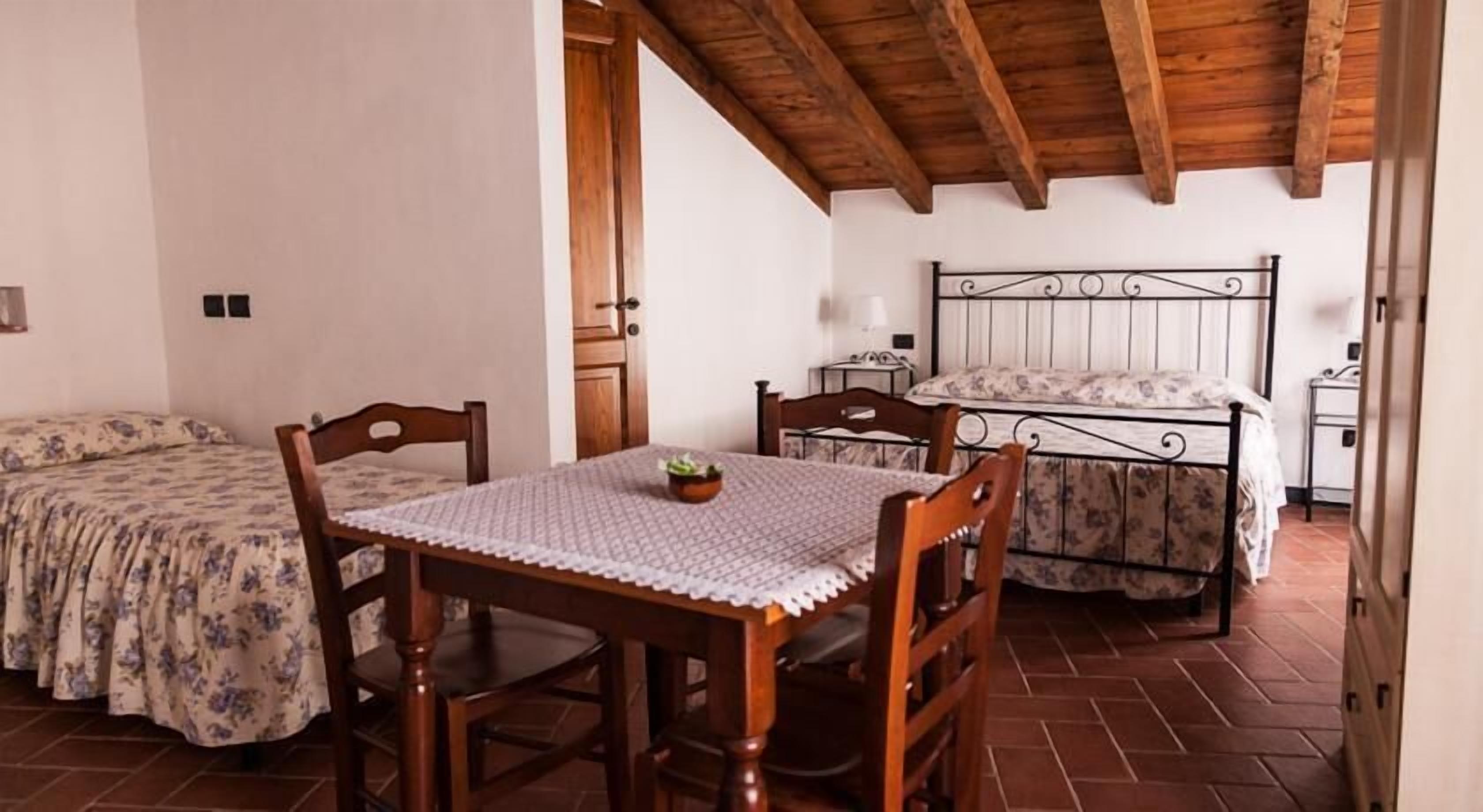 A bedroom with a wooden ceiling, a bed with a floral bedspread, a bedside table with a lamp, and a small table with chairs in the center.
