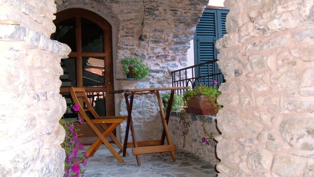 Un espacio al aire libre con paredes de piedra, una mesa y sillas de madera, una planta en maceta y vistas a un edificio con contraventanas azules.