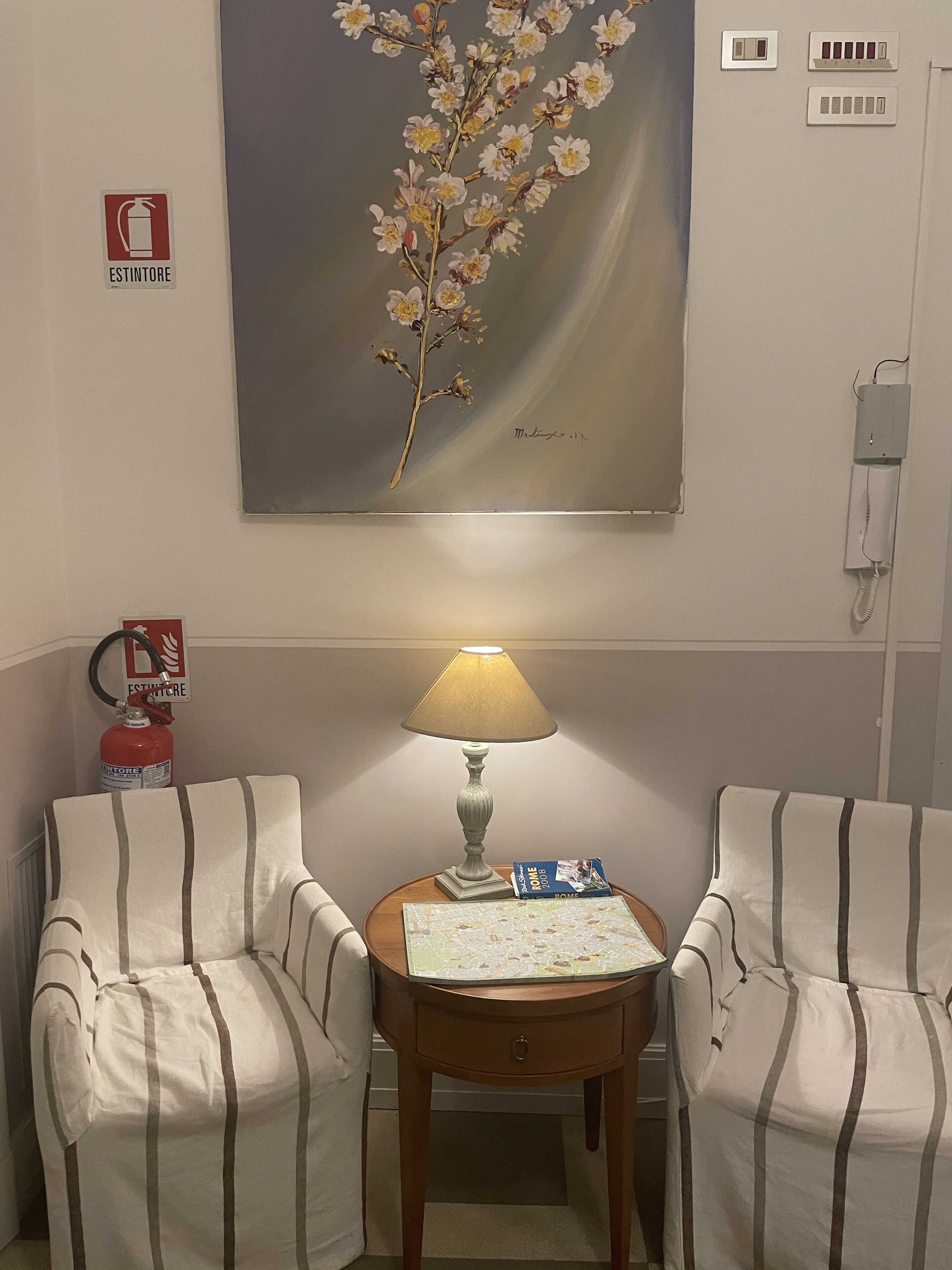 Ein Zimmer mit zwei gestreiften Sesseln, einem Tisch mit Lampe und Buch, einem Blumenbild, einem Feuerlöscher und einem Telefon an der Wand.