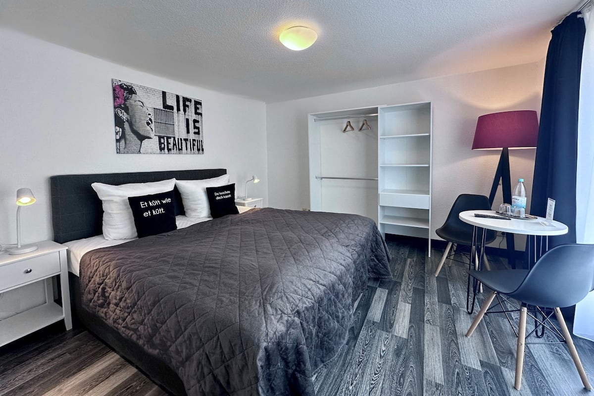 A modern hotel room with a large bed, a wardrobe, a desk with a chair, and a wall with a motivational quote.