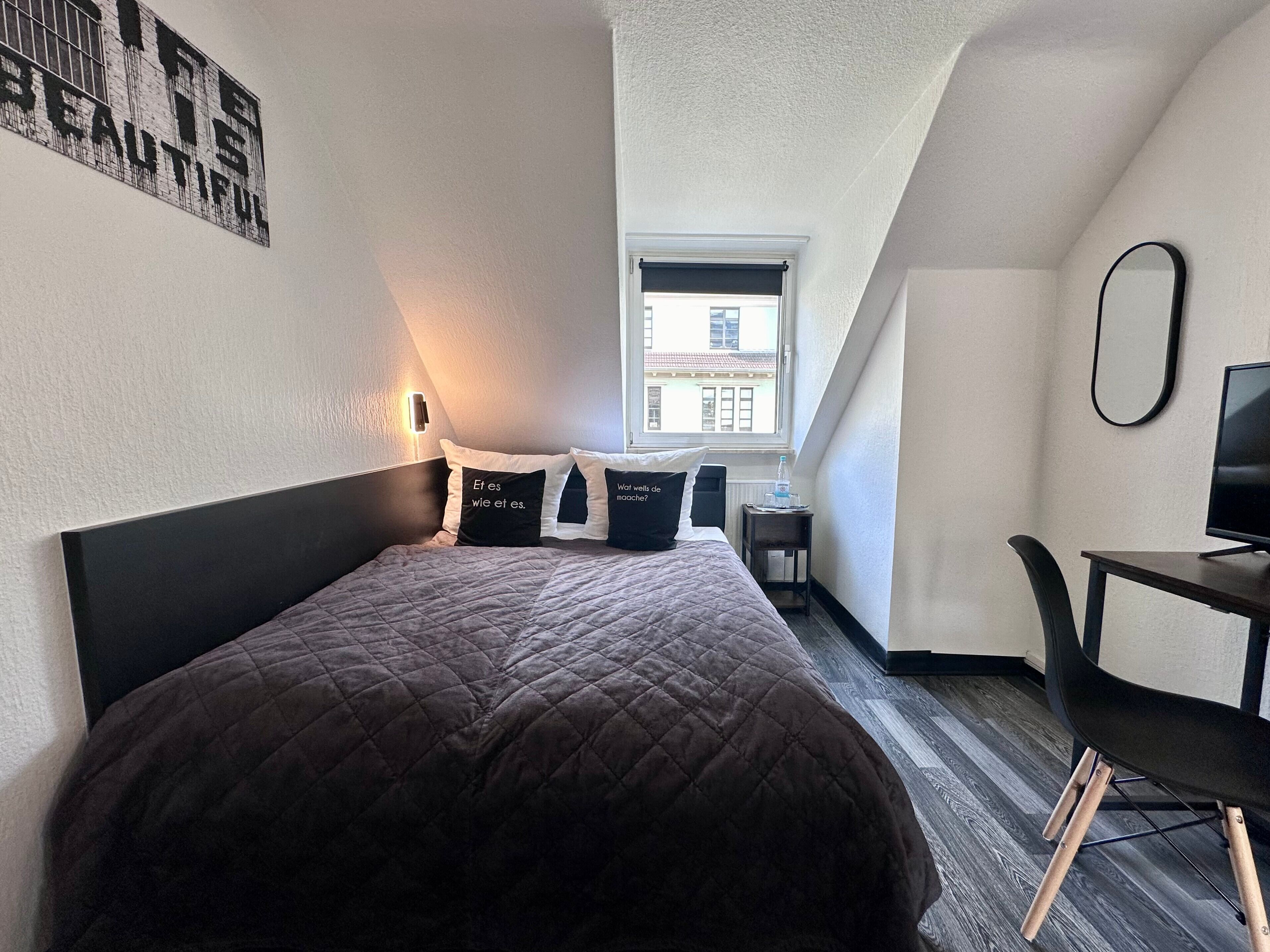 Une chambre à coucher comprenant un lit, des oreillers, un bureau, une chaise, un téléviseur et une fenêtre donnant sur des immeubles.
