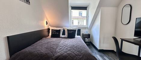 Une chambre à coucher comprenant un lit, des oreillers, un bureau, une chaise, un téléviseur et une fenêtre donnant sur des immeubles.