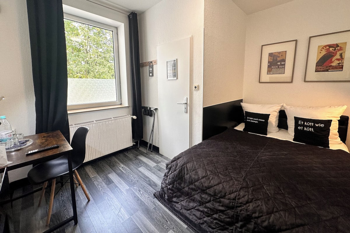 A hotel room with a bed, a desk with a chair, a radiator, a window with curtains, and framed pictures on the wall.