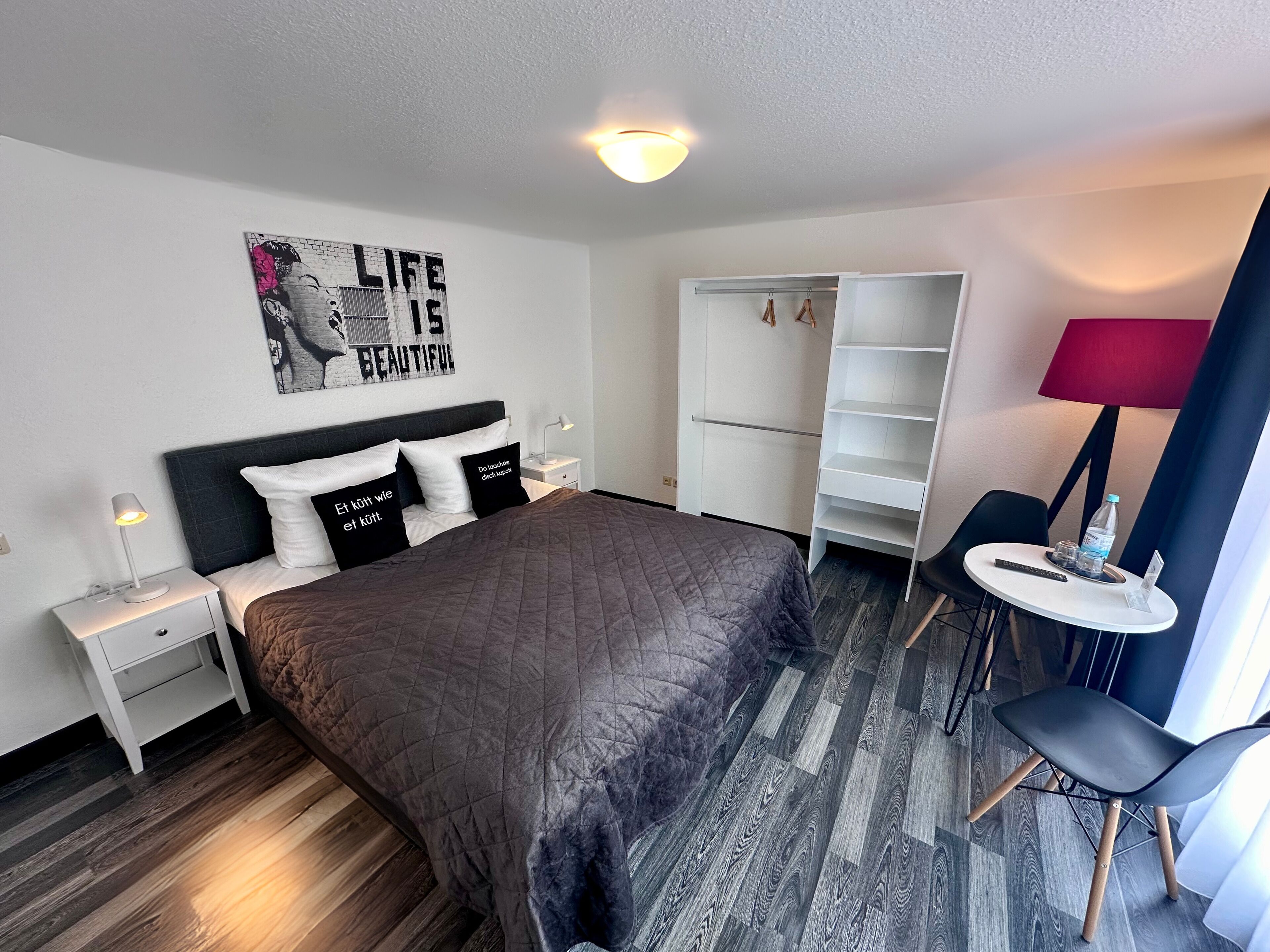 A hotel room with a bed, a nightstand, a desk, a chair, a wardrobe, and a small table with a lamp.