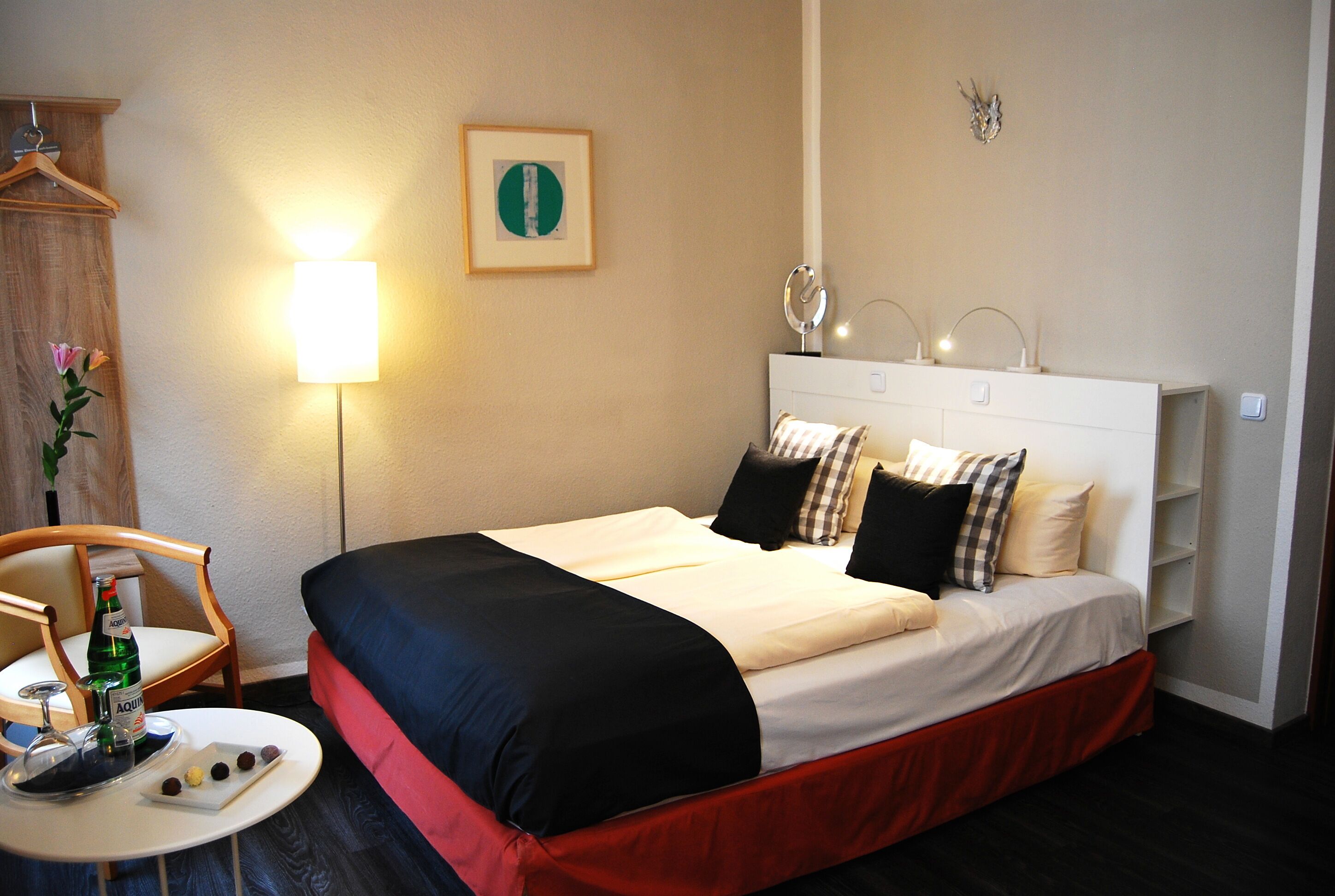 A neatly made bed with a red skirt, a bedside table with a bottle and glasses, and a hanging plant.