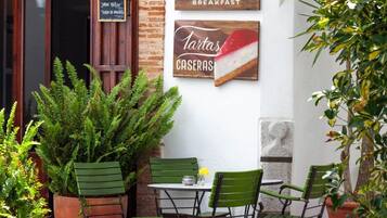 Un área de estar al aire libre con sillas verdes y una mesa, rodeada de plantas en macetas y una pared de ladrillo.