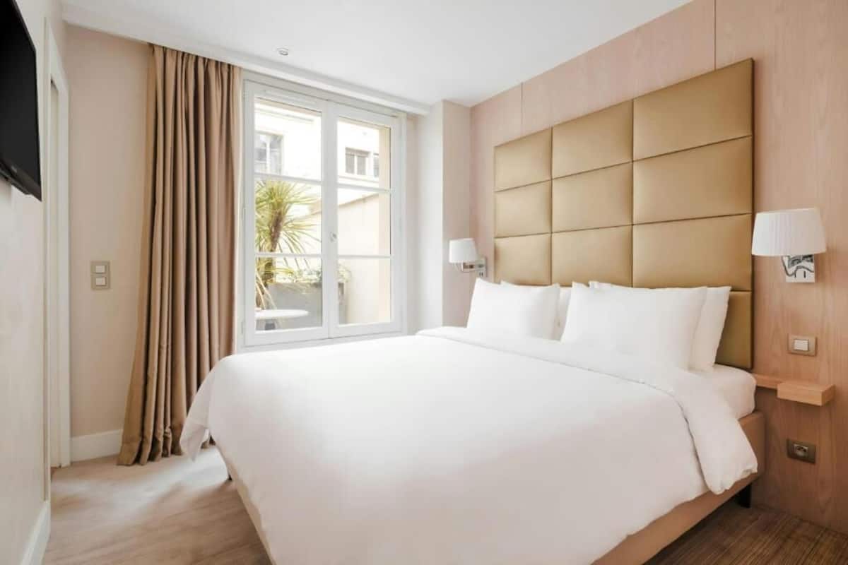 A hotel room with a large bed, a window with curtains, a wall-mounted TV, and a bedside table with a lamp.