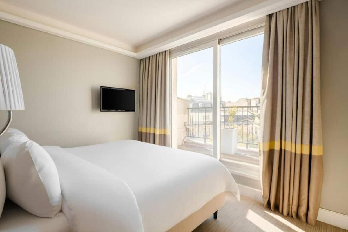 A hotel room with a large bed, a flat-screen TV mounted on the wall, and a balcony with curtains.