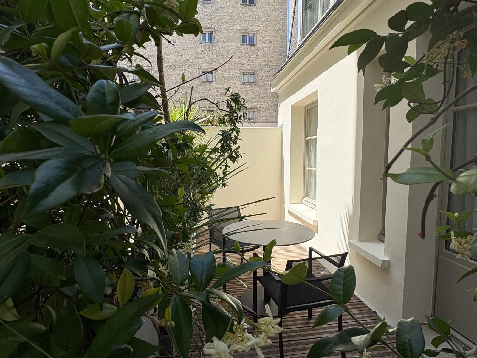 A small outdoor seating area with a table and chairs, surrounded by lush greenery and a building in the background.