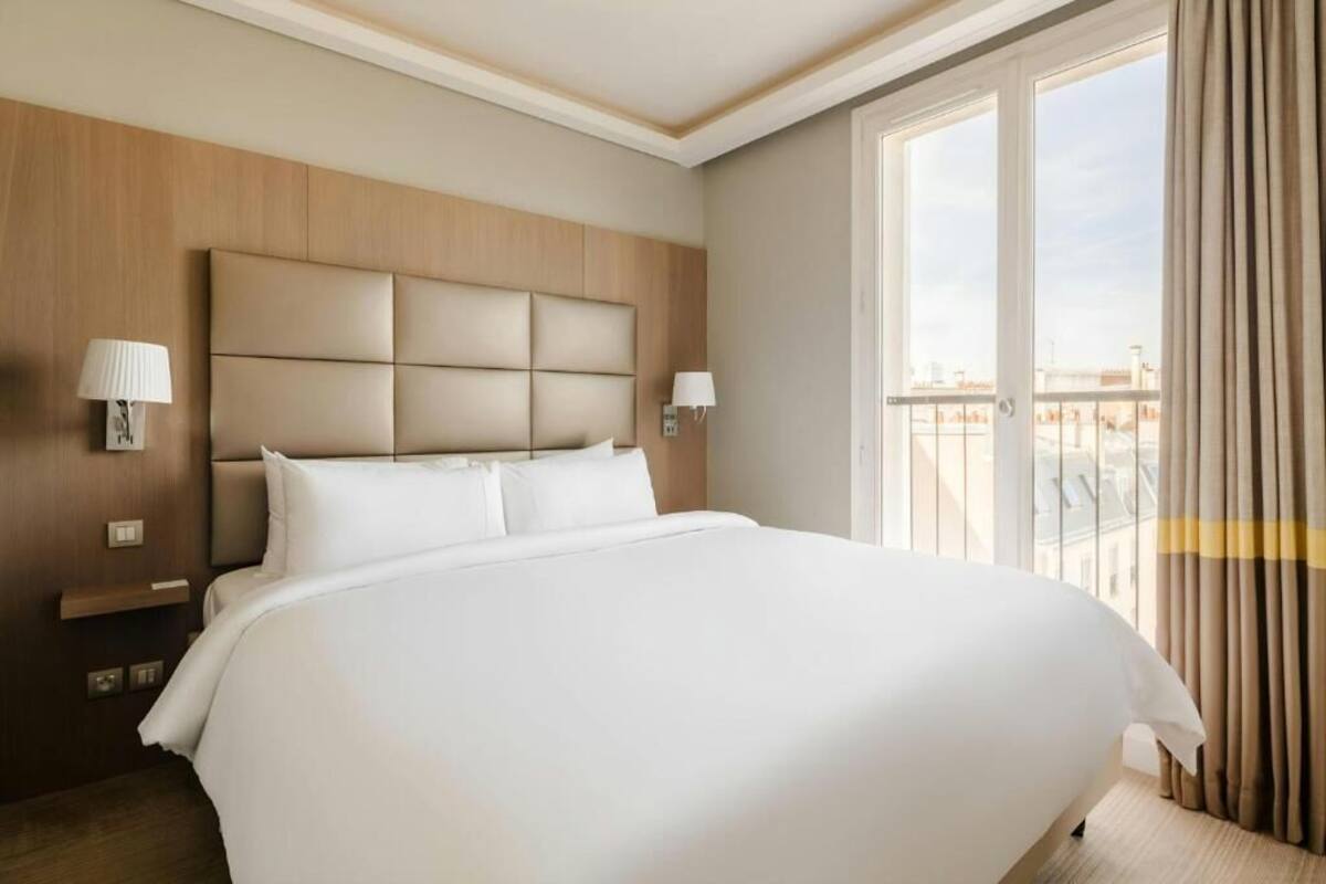 A large bed with a wooden headboard, two bedside lamps, and a view of the city through a balcony with curtains.