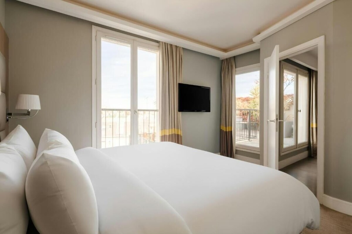 A hotel room with a large bed, a flat-screen TV mounted on the wall, and a balcony with curtains.