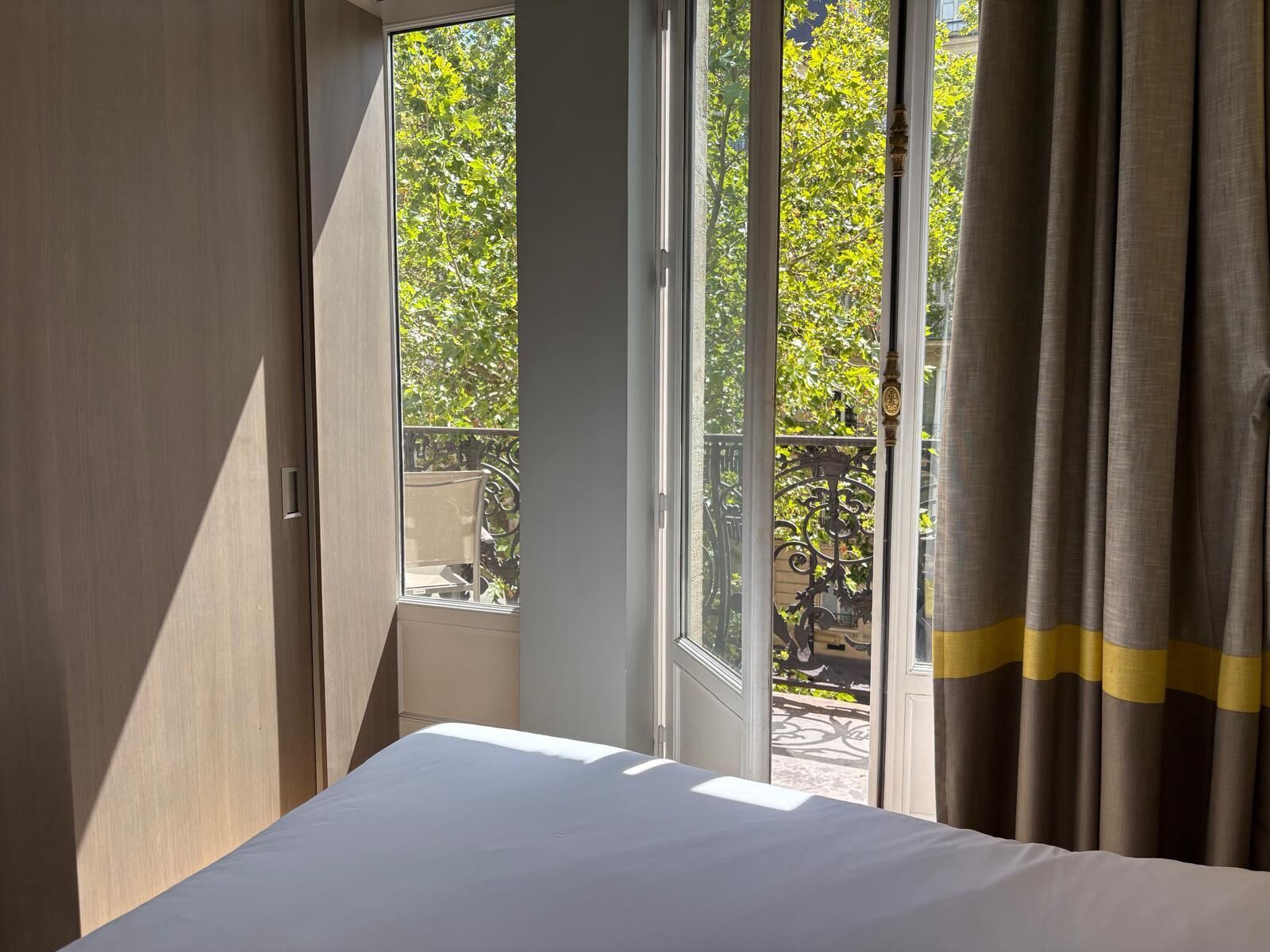 Una habitación de hotel con cama, ventana con cortinas y vistas a un balcón con mobiliario exterior.