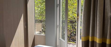 Una habitación de hotel con cama, ventana con cortinas y vistas a un balcón con mobiliario exterior.