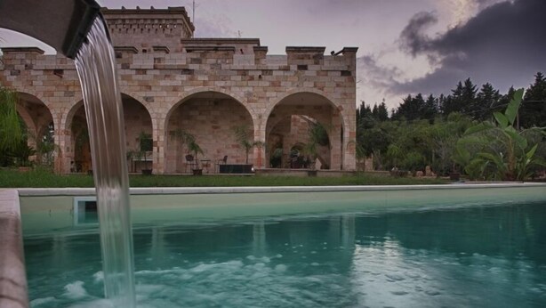 A pool with a waterfall feature and a stone building with arches in the background. - Relais Villa Lucia (Carovigno)