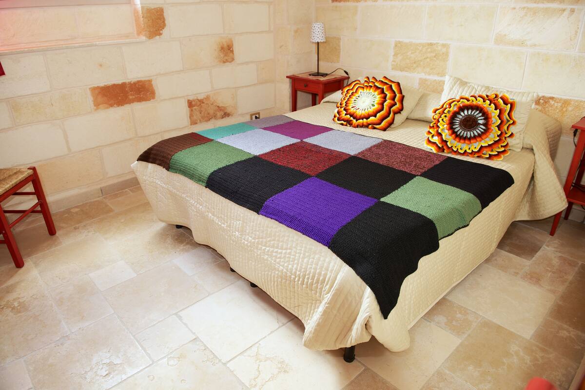 Uma cama bem arrumada com um colcha colorida, uma pequena mesa de cabeceira com abajur e uma cadeira em um quarto com piso de azulejos e paredes de pedra.