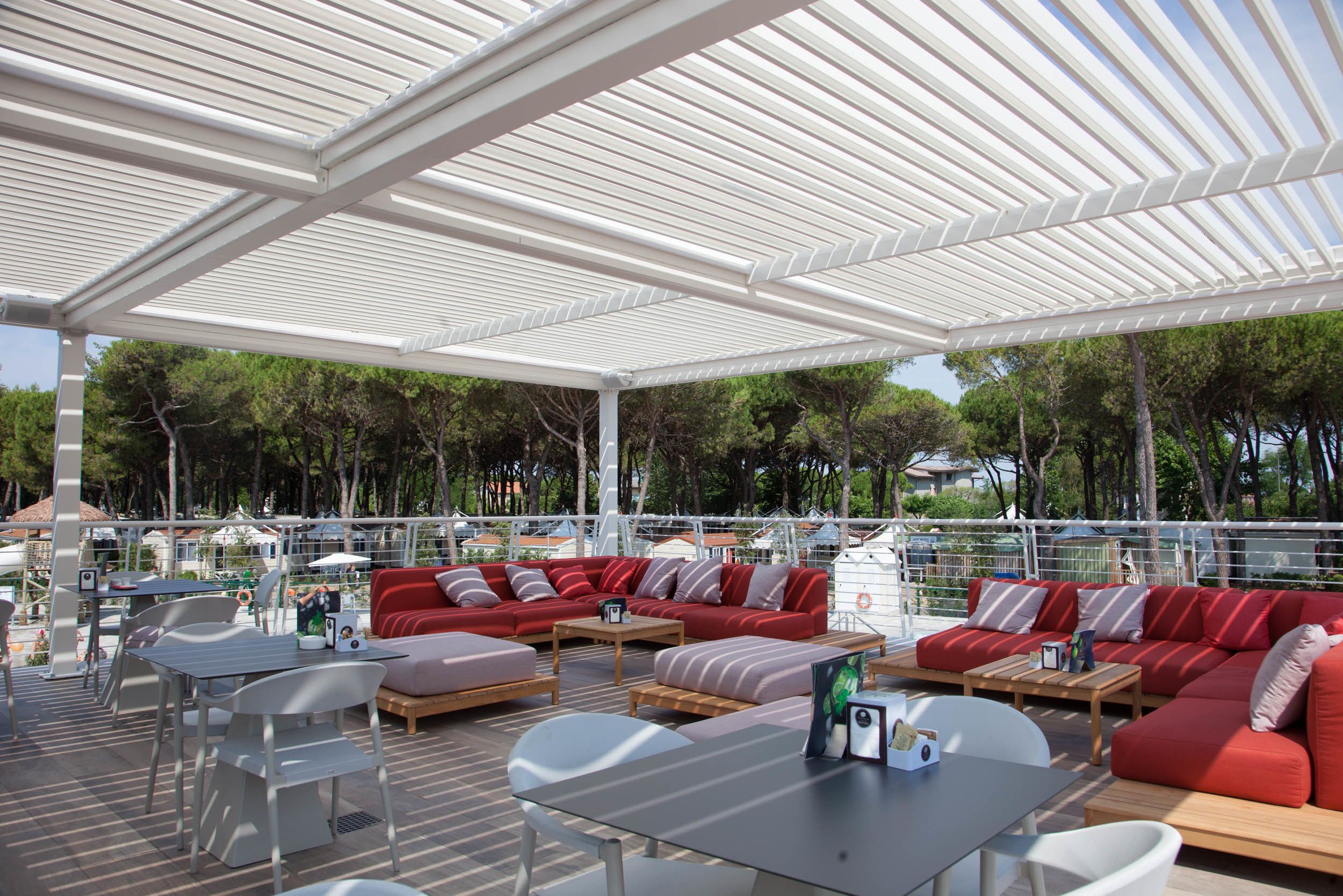 An outdoor seating area with red sofas and white tables, surrounded by a white pergola.