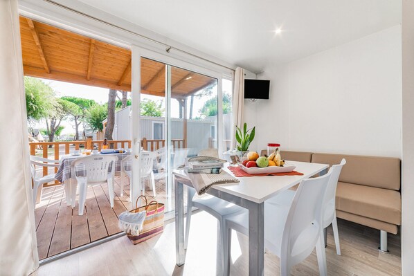 A modern outdoor dining area with a wooden deck, white plastic chairs, a table with a fruit bowl, and a beige sofa. - Camping Village Cavallino (Cavallino-Treporti)