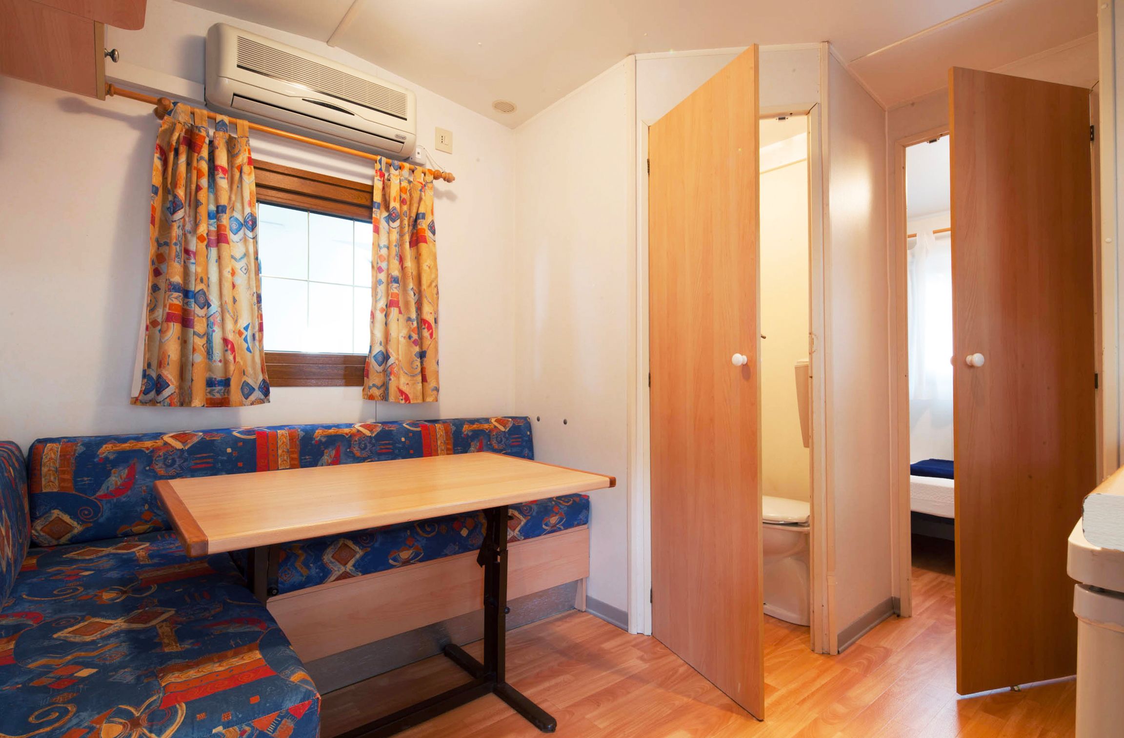 Een kleine kamer met een houten tafel, een bank met gestreept bekleding, een raam met gordijnen, en een deur naar een andere kamer.