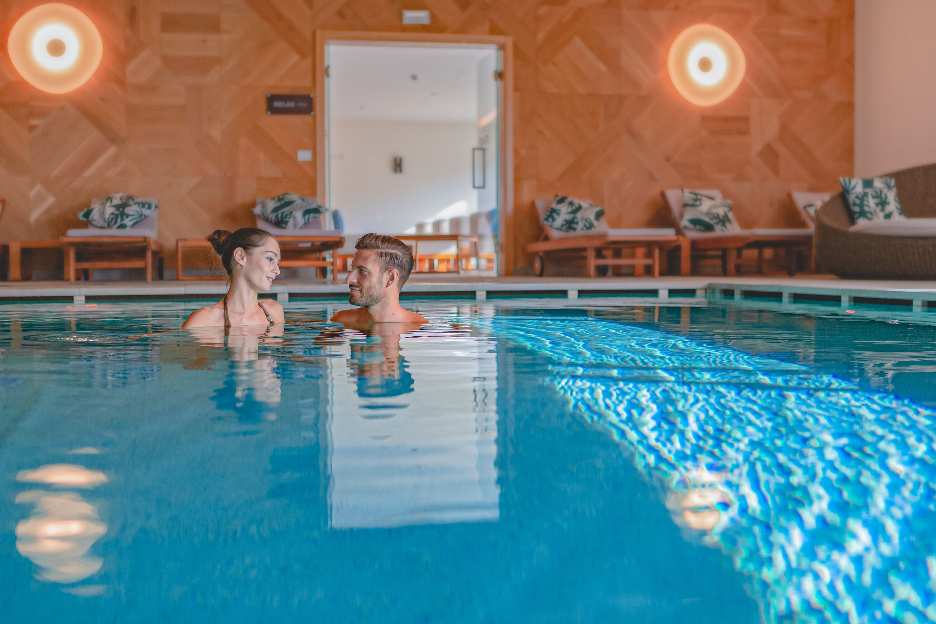 Une piscine où se trouvent deux personnes, entourée de chaises longues et de murs en bois.