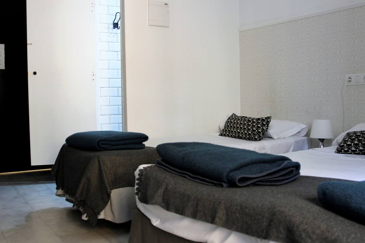 A hotel room with two beds, each with a dark-colored bedspread and pillows, a bedside table with a lamp, and a window with a view of the outside.