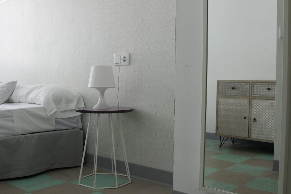 A bedroom with a bed, a small table with a lamp, a wardrobe, and a patterned wall.