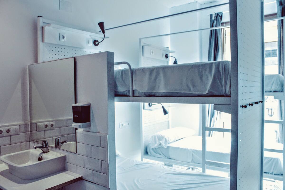 A bunk bed room with a sink and mirror, a window with curtains, and a wall-mounted light fixture.