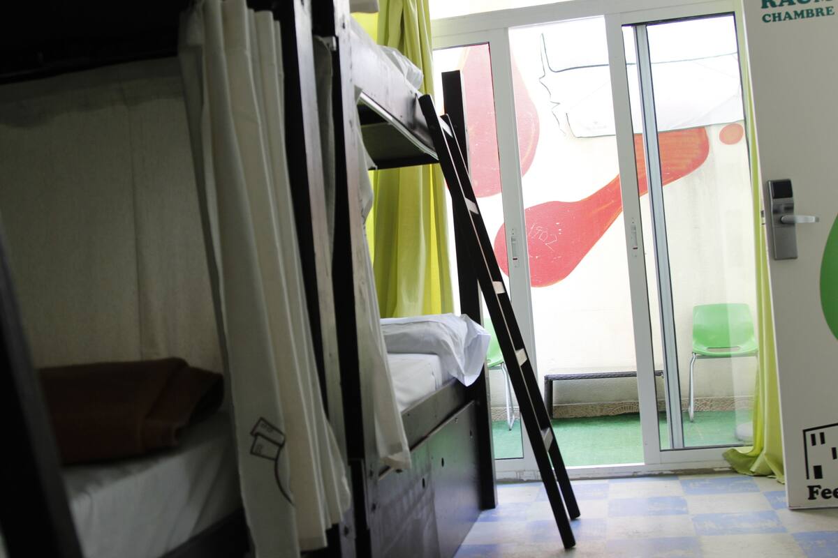 A dormitory room with bunk beds, a sliding glass door leading to a balcony, and a green chair outside.