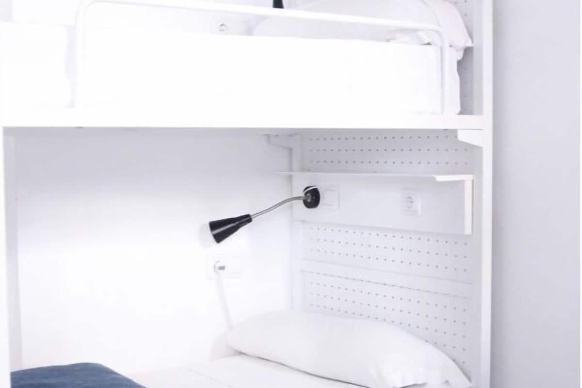 A white, compact room with a bed, a small bedside table, and a wall-mounted lamp.