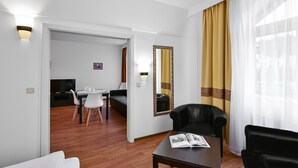 Een moderne hotelkamer met een houten vloer, een zithoek met een tafel en stoelen, een zwarte leren bank, een klein tafeltje met een tijdschrift, en een grote spiegel met een decoratieve lijst.