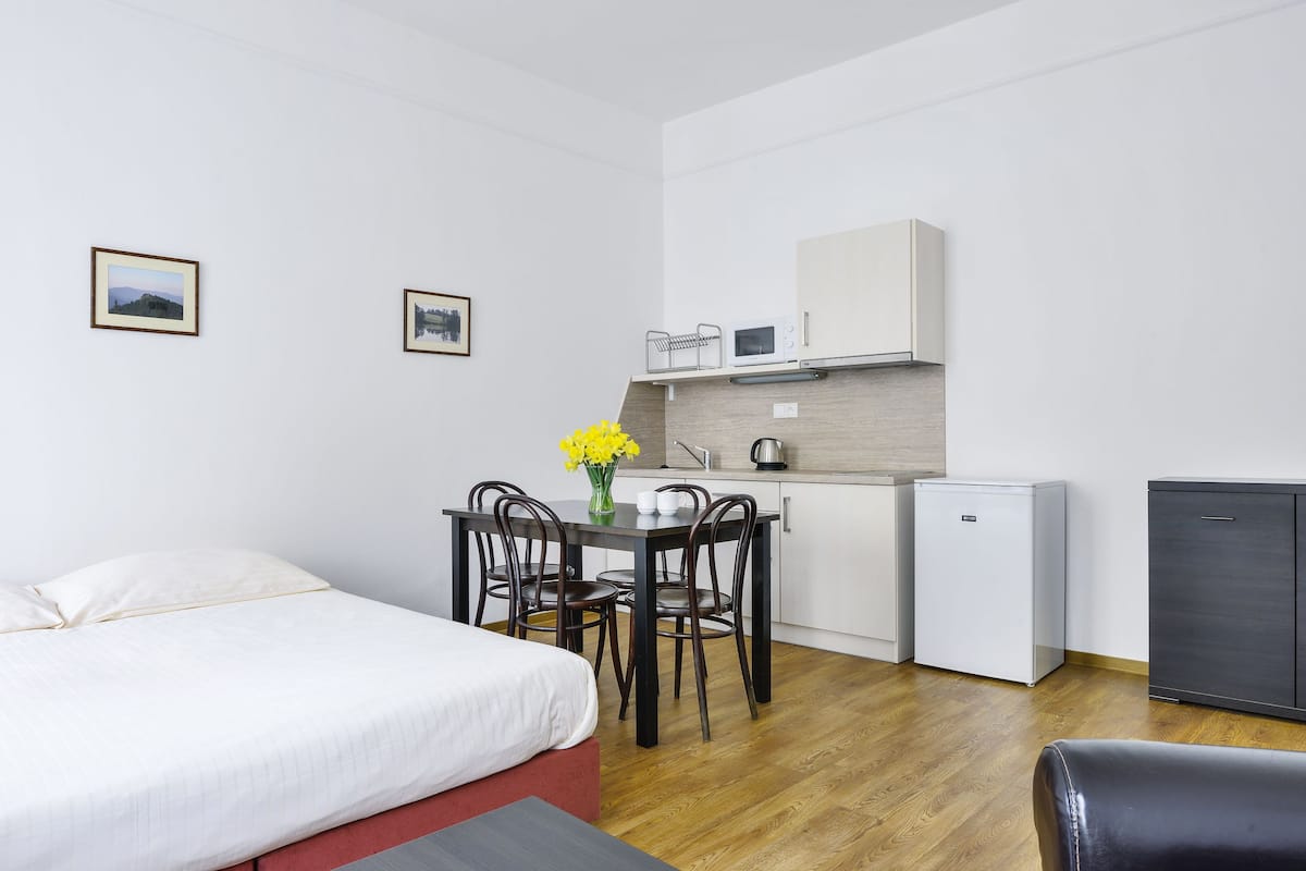 A hotel room with a bed, a small dining table with chairs, a kitchenette, and a compact refrigerator.