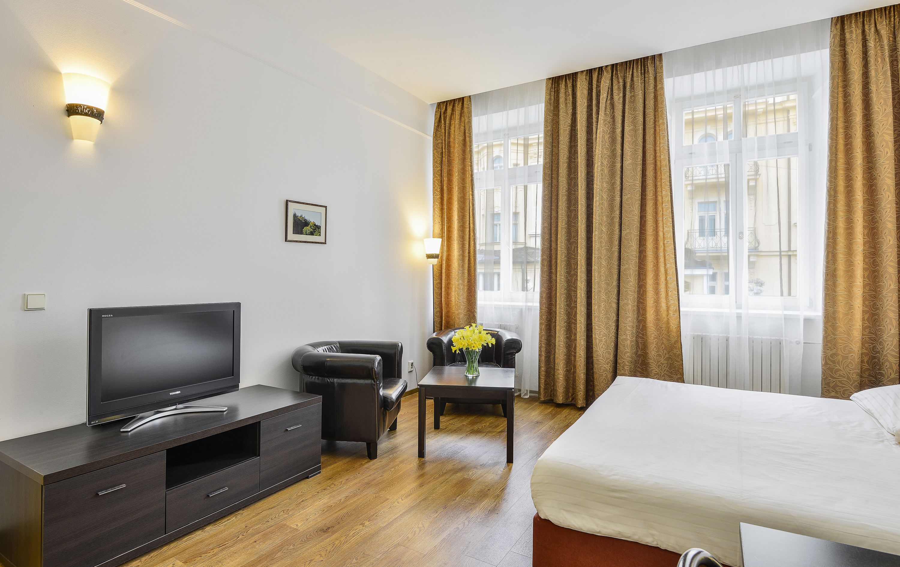 A hotel room with a bed, a TV, a desk with a vase of flowers, and a chair.