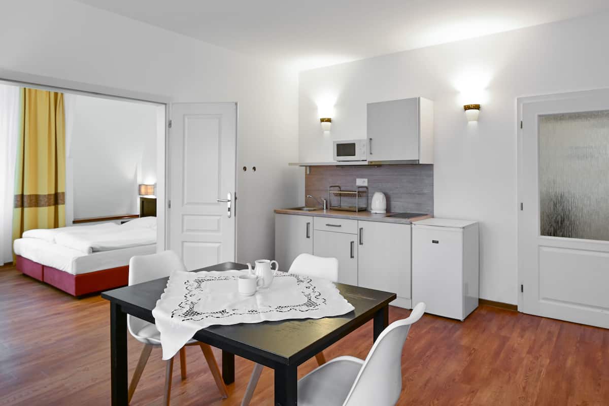 A compact hotel room with a bed, a small kitchenette, a dining table with chairs, and a view of the city through a window.