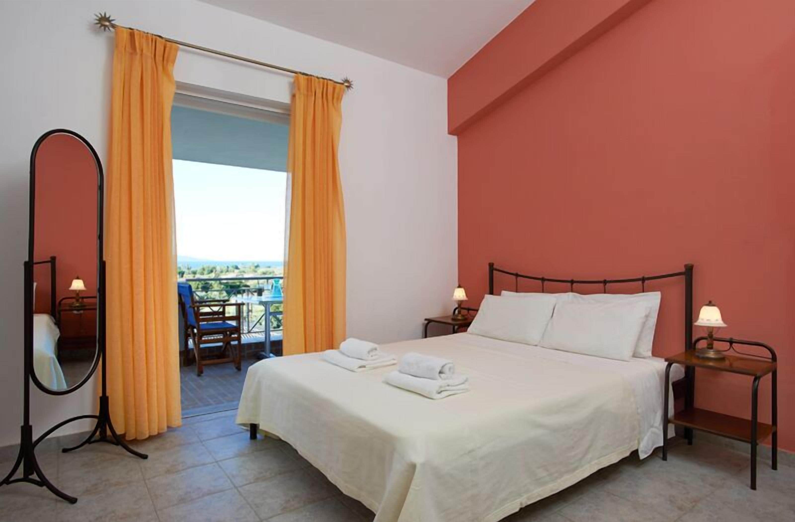 Quarto de hotel com cama, duas mesas de cabeceira com luminárias, espelho de corpo inteiro e varanda com cortinas laranja.