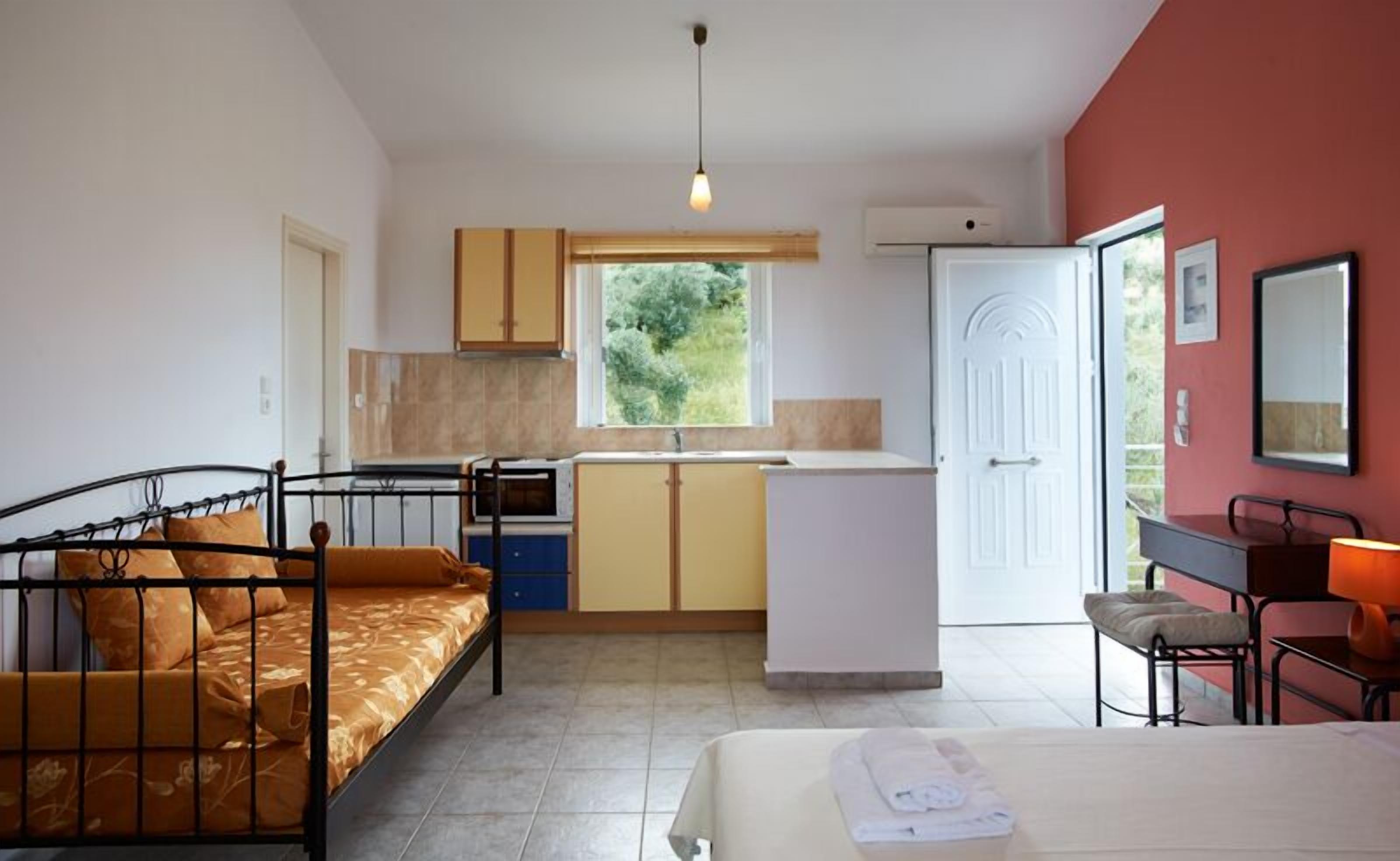 A compact room with a bed, a small kitchenette, a window with a view of greenery, and a door leading to an outdoor area.