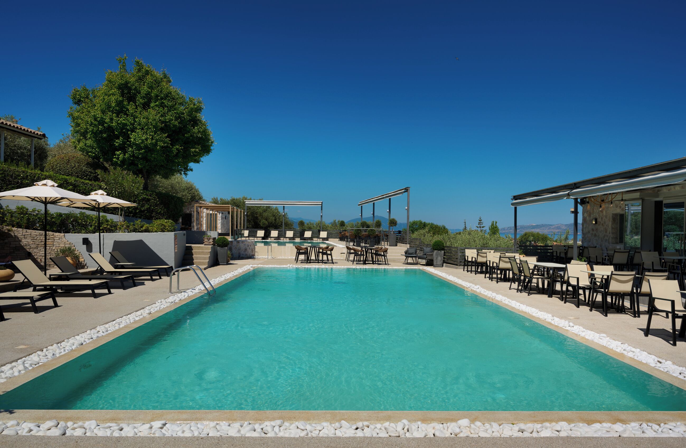 Piscina azul límpida com espreguiçadeiras e guarda-sóis, cercada por um muro de pedra e vegetação.