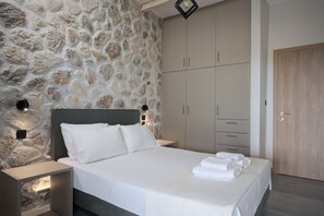 A bedroom with a stone wall, a bed with white linens, a wooden wardrobe, and a nightstand with a lamp. - Irene Studios (Istiaia-Aidipsos)