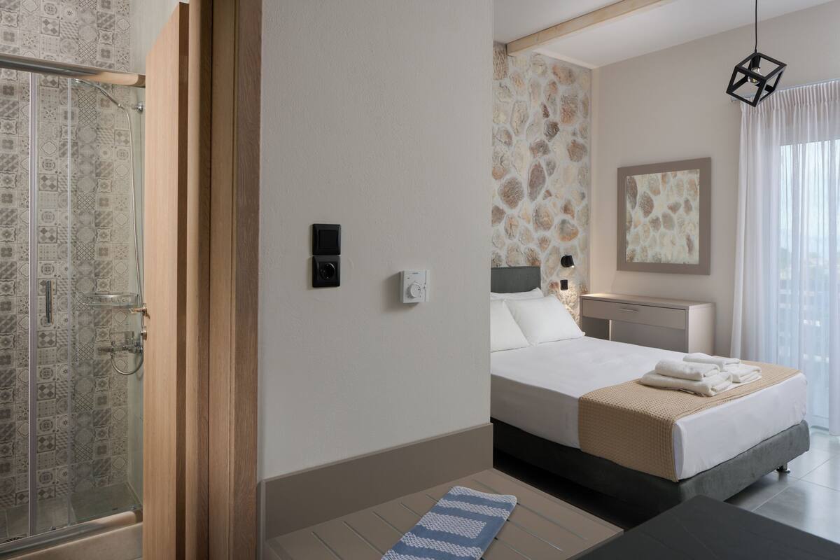 A hotel room with a bed, a nightstand, a mirror, a window with curtains, and a shower area with a patterned tile wall.